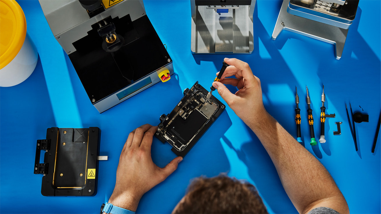 Homme répare un téléphone sur une table bleue avec un tourne vis et différents outils et pièces autour