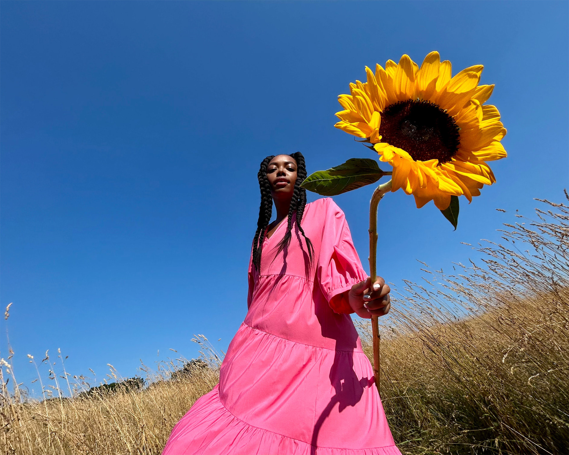 Femme dans une robe rose prise en contre plongée tenant un tournesol dans un champ de blé avec un ciel bleu en arrière plan