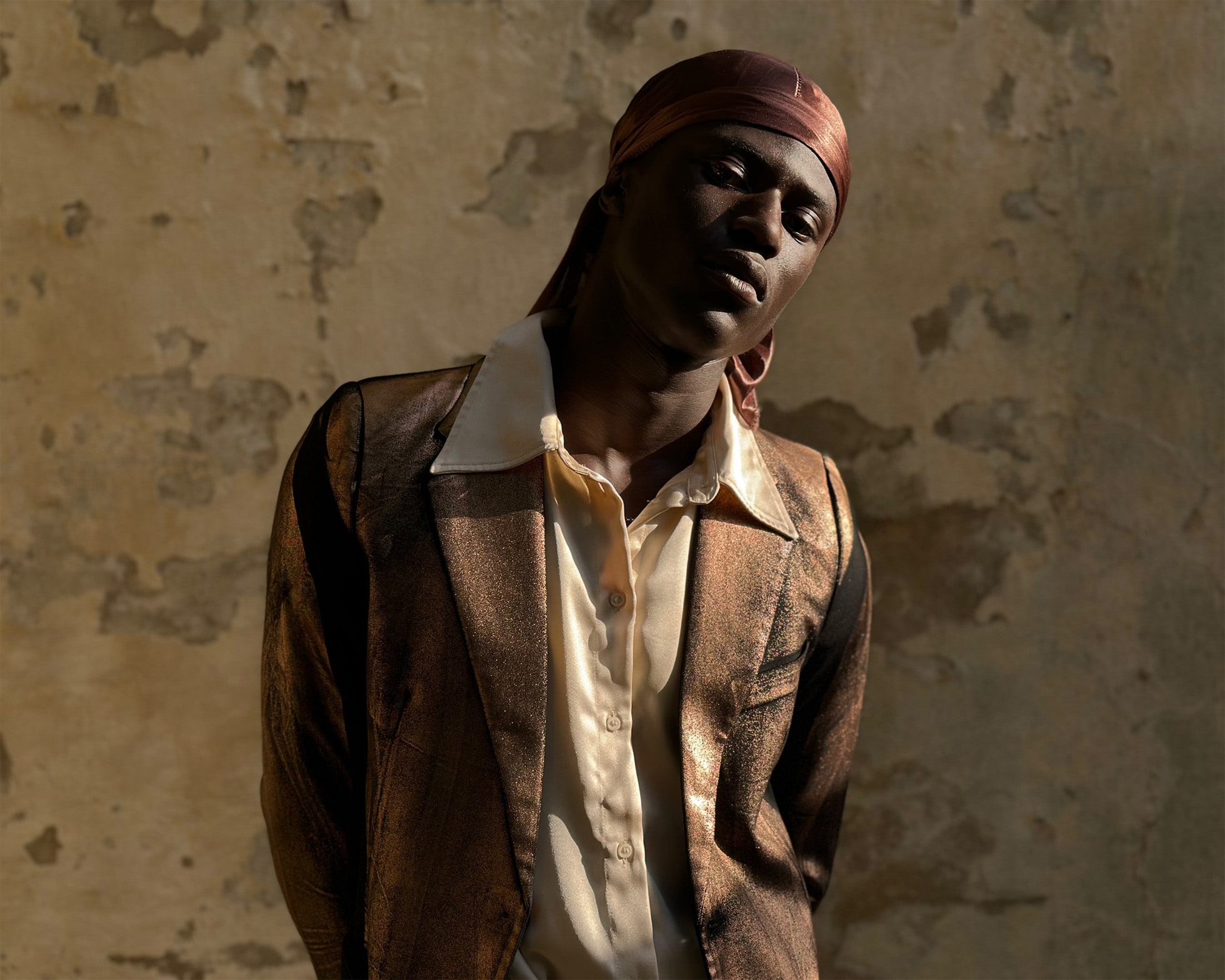 Photo du buste d'un homme portant une veste de costume dorée et une chemise blanche dans une pièce vide, il regarde l'objectif en penchant la tête et porte une boucle d'oreille à l'oreille gauche