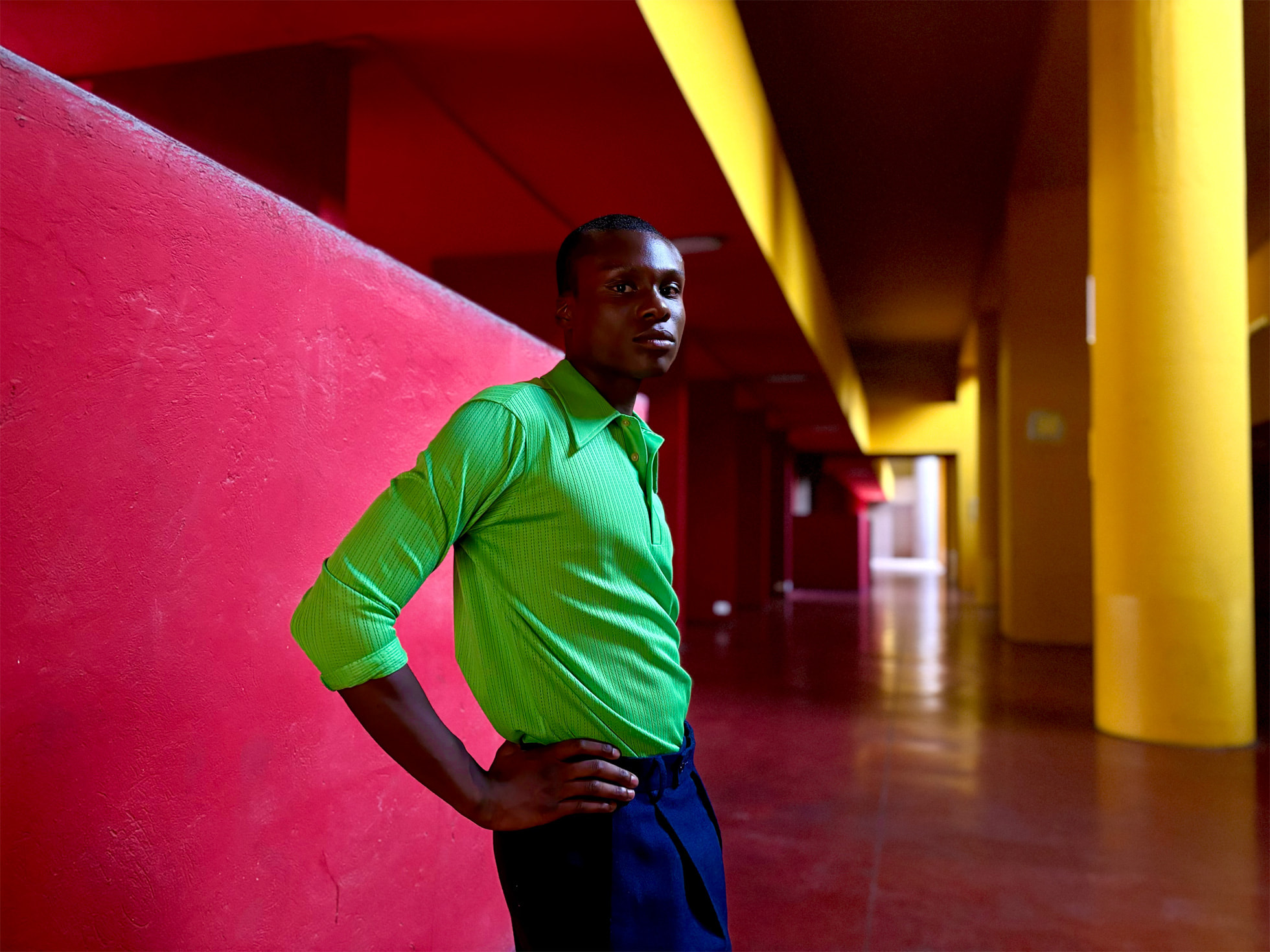 Homme dans un long couloir coloré de rose et de poutres jaunes, vêtu d'une chemise verte unie, nous regardant dans les yeux