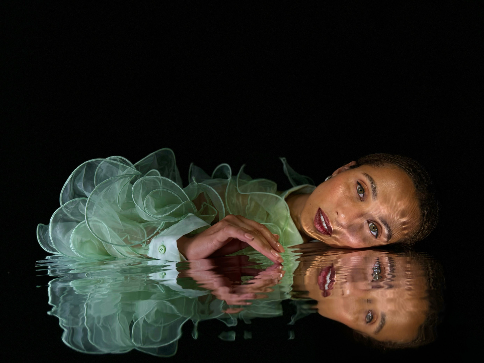 Femme posant avec élégance, vêtue d'une robe en tissu translucide vert, allongée sur une surface d'eau réfléchissante avec un éclairage sombre et dramatique, mettant en valeur ses traits et son regard intense.