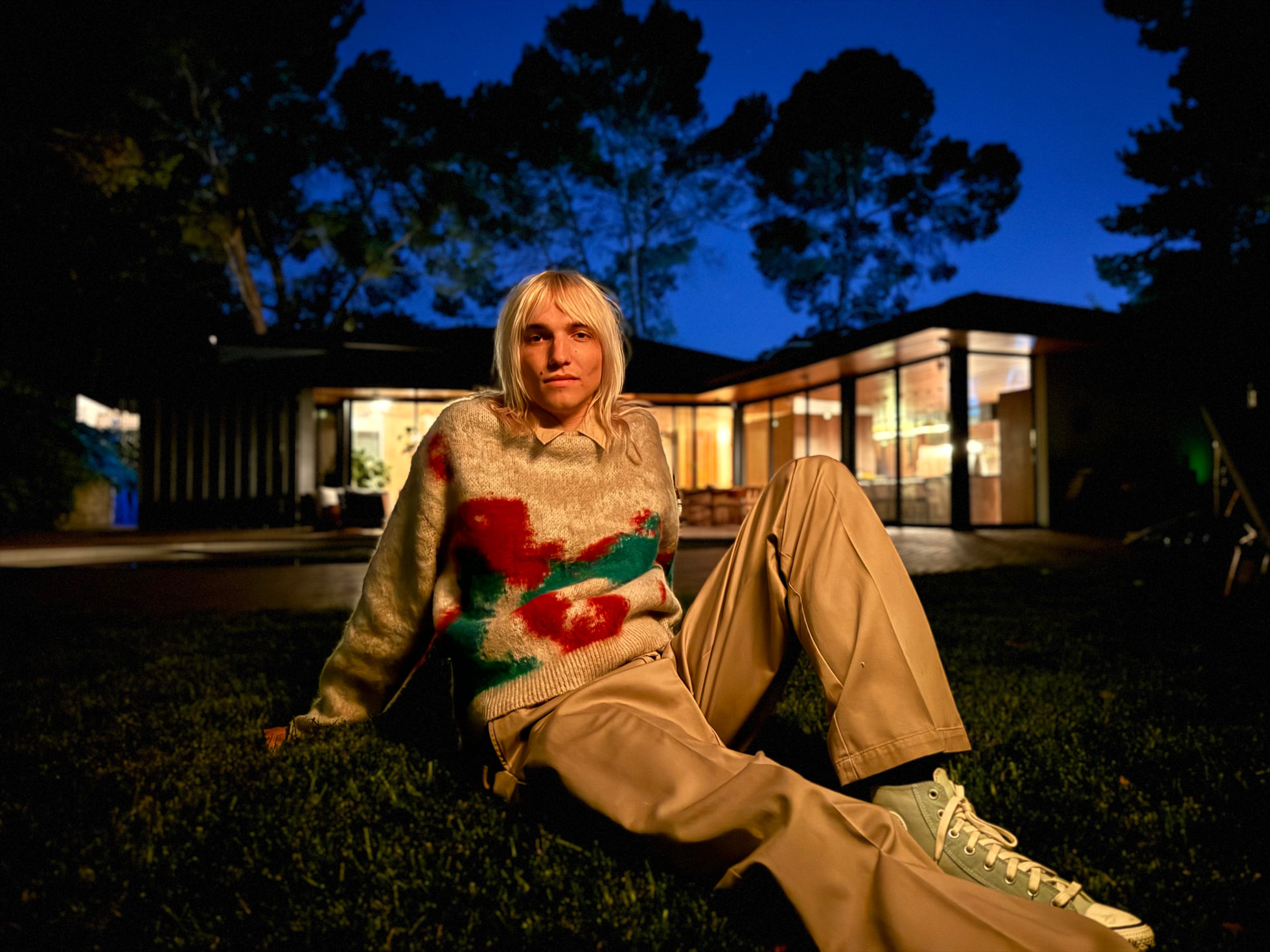 Personne assise sur une pelouse la nuit, portant un pull coloré et un pantalon beige, avec une maison moderne éclairée en arrière-plan et un ciel bleu profond.