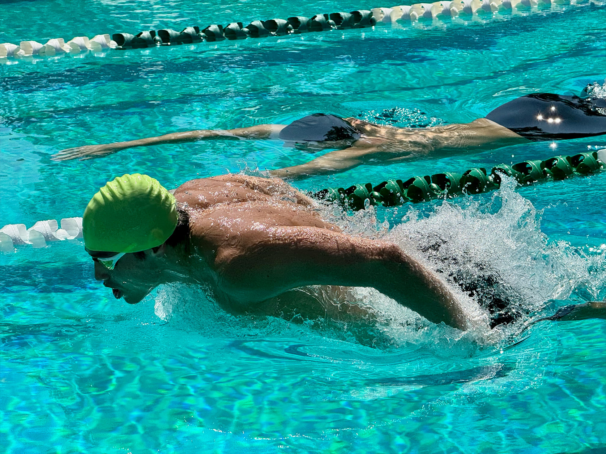 Nageur portant un bonnet de bain vert et des lunettes, exécutant le papillon dans une piscine turquoise, avec un autre nageur en arrière-plan dans une autre ligne d'eau.