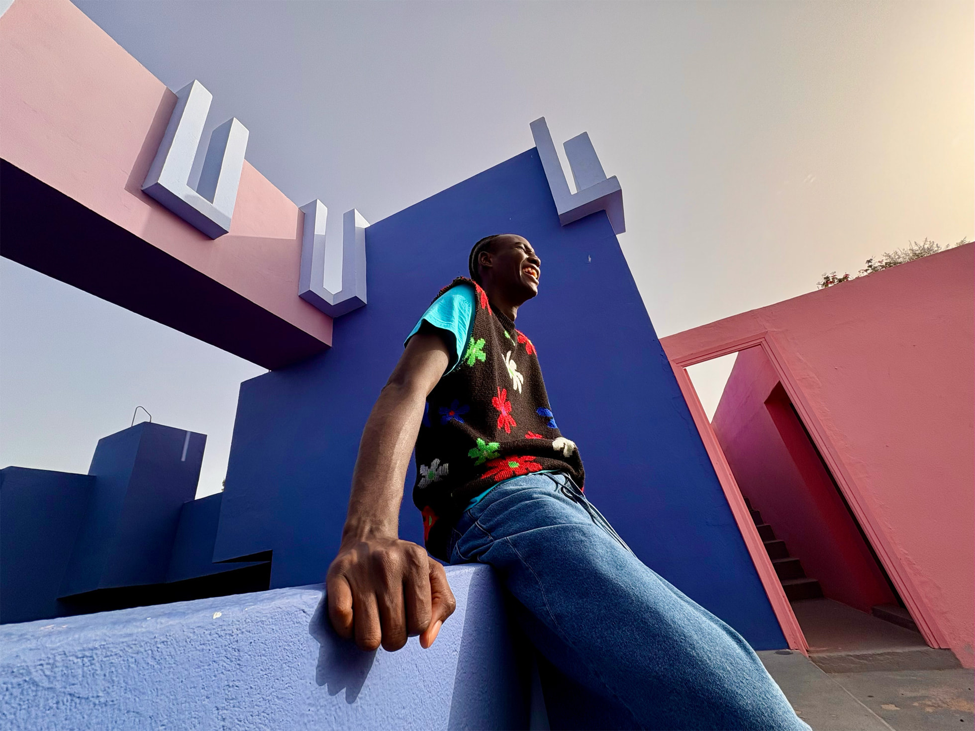 Une image d’un homme souriant en vêtements colorés s’appuyant sur un bâtiment rose et mauve.