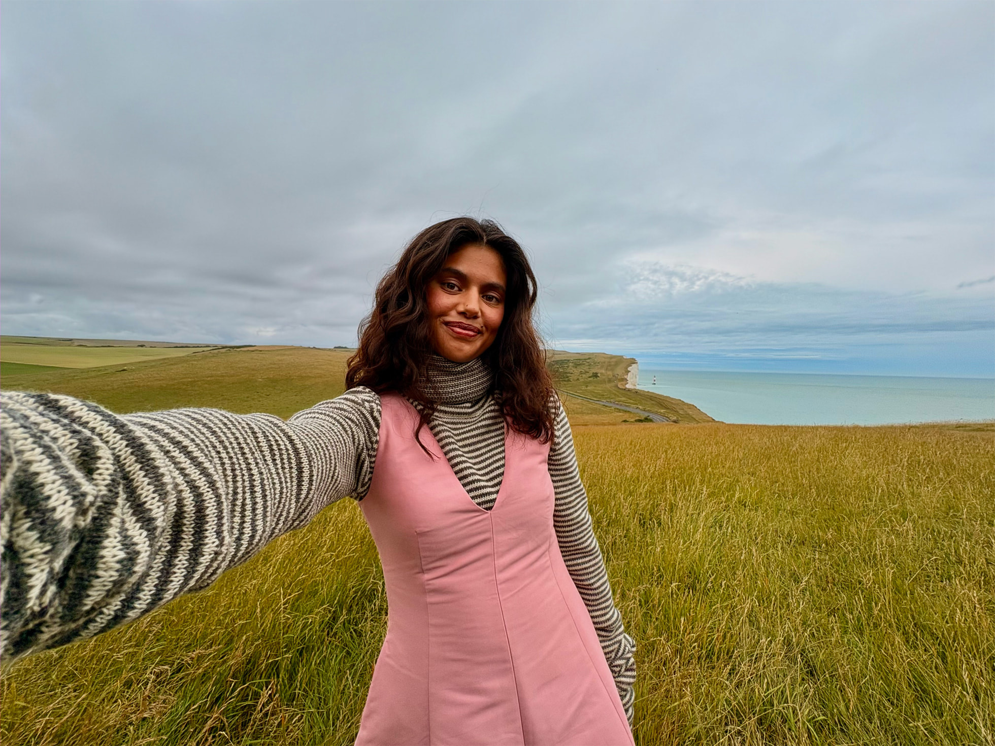 Un selfie d'une femme en robe rose avec un sous t-shirt zébré avec en arrière-plan un paysage verdoyant menant à la mer.